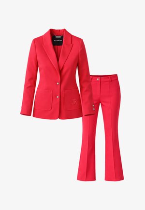 Completo donna su misura rosso con blazer aderente caratterizzato da due bottoni argento, tasche frontali e pantaloni svasati abbinati con chiusura a bottone argento.