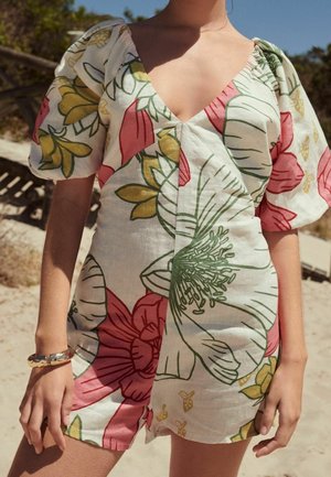 Femme portant une robe courte à col en V avec des manches bouffantes et de grands imprimés floraux roses, verts et jaunes à l'extérieur par une journée ensoleillée.