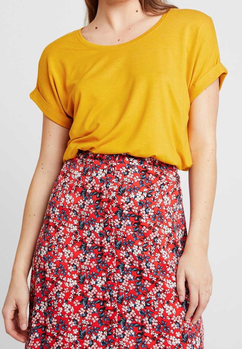 T-shirt giallo a maniche corte e collo rotondo abbinato a una gonna rossa con pattern floreale, caratterizzata da fiori bianchi e blu su uno sfondo vivace.