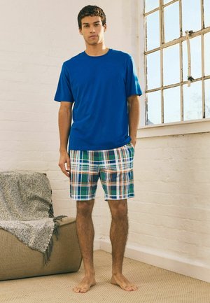 Man standing barefoot indoors wearing a blue short-sleeve shirt and green, white, and orange plaid shorts by a window and sofa.