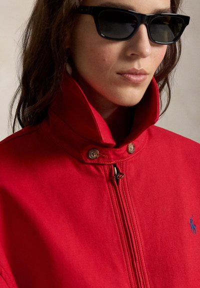 Femme portant des lunettes de soleil rectangulaires noires et une veste zippée rouge à col montant avec un logo bleu brodé.