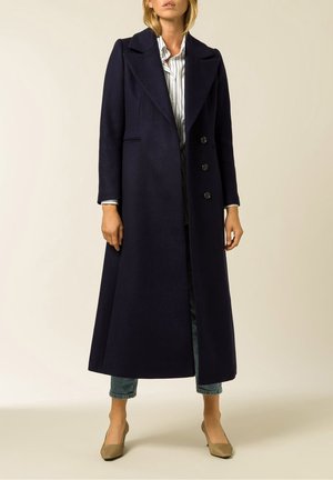 Frau trägt langen marineblauen Mantel mit weitem Revers, weiß gestreiftes Hemd, gekürzte Jeans und beige Absätze vor hellem Hintergrund.