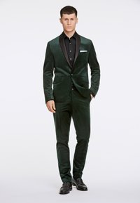 Sacco di velluto verde scuro con revers in raso nero, abbinato a una camicia nera a bottoni, pantaloni slim fit e scarpe nere. Texture liscia.