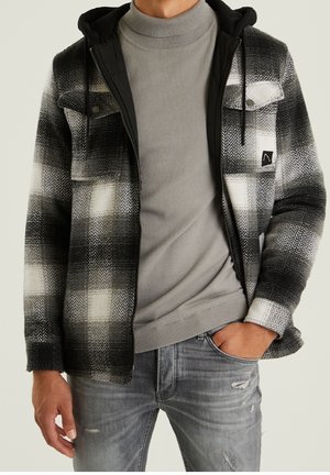 Hombre con chaqueta de cuadros en blanco y negro con cremallera y capucha, suéter de cuello alto gris y jeans grises desgastados, posando con una mano en el bolsillo.