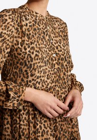 Vestido de estampado de leopardo en tejido ligero, con cuello tipo camisa, detalles de botones y puños fruncidos. Patrón marrón y negro sobre fondo beige.