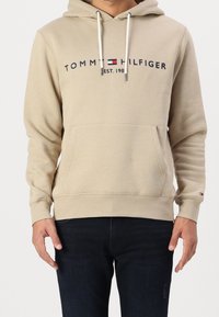Tan hooded sweatshirt with a front pocket, featuring a black logo and red accents. Soft texture and relaxed fit.