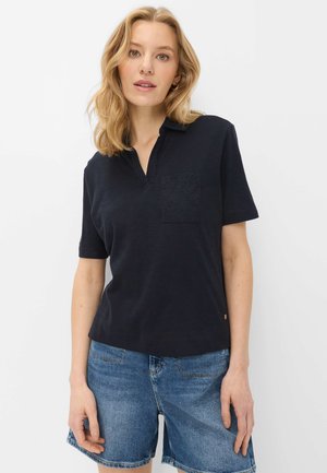 Blonde vrouw draagt een zwart kort mouws overhemd met kraag en een kanten zakje, gecombineerd met blauwe denim shorts, staand tegen een eenvoudige achtergrond.