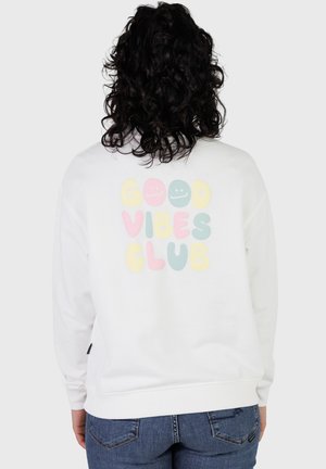 Person mit lockigem schwarzen Haar, die ein weißes Sweatshirt mit pastellfarbenem "GOOD VIBES CLUB"-Schriftzug und lachenden Gesichtern auf dem Rücken trägt, kombiniert mit blauer Jeans.