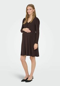 Zwangere jurk in donkerbruin ribbelstof met lange mouwen, V-hals en knoopdetails. Gladde textuur en knielange afwerking met een ontspannen pasvorm.