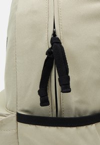 Mochila de tela beige con una superficie texturizada, que cuenta con una cremallera y un tira negra. Incluye una correa negra alrededor de la base para mayor soporte.