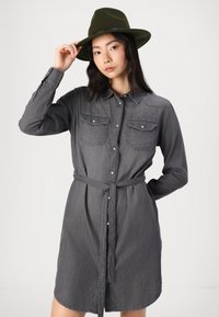 Robe chemise en denim gris avec fermeture à boutons, col et deux poches poitrine ; taille cintrée avec une ceinture assortie ; portée avec un chapeau vert foncé.