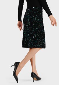 Falda negra con lentejuelas y acentos verdes brillantes, de longitud hasta la rodilla, combinada con tacones altos negros puntiagudos. Textura suave y diseño ajustado.