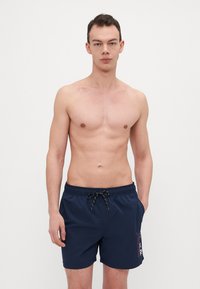 Marineblå svøm shorts med snøring i livet og sidelommer. Har et grafisk trykk på venstre ben. Laget av lett og hurtigtørkende materiale.