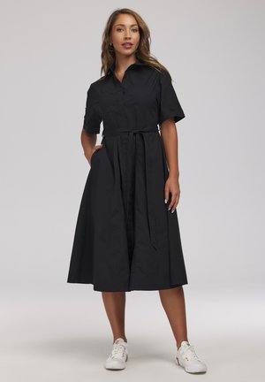 Kobieta w czarnej midi sukience na guziki z krótkim rękawem i wiązaniem w talii, stojąca z jedną ręką w kieszeni, białe trampki, neutralne tło.