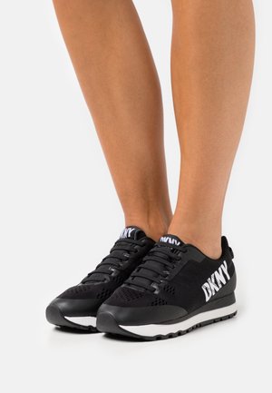 Schwarze Sportschuhe mit Mesh-Obermaterial, verstärkter Zehenpartie, weißem DKNY-Logo an der Seite und kontrastierender weißer Sohle.