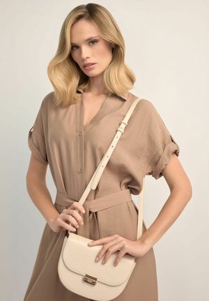 Blondynka w beżowej sukience z paskiem, trzymająca kremową torebkę na ramię z złotym zapięciem na neutralnym tle.