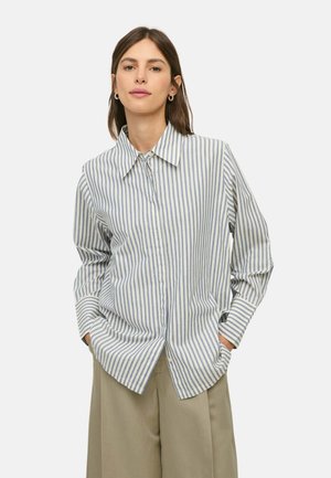 Mujer con una camisa azul y blanca de botones con rayas verticales holgada y pantalones anchos beige, de pie con las manos en los bolsillos.