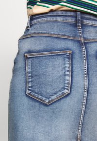 Blå denimkjol med en bakficka med en sydd kant, medelhavs-tvätt och slät yta. Framträdande med kontrasterande sömmar.