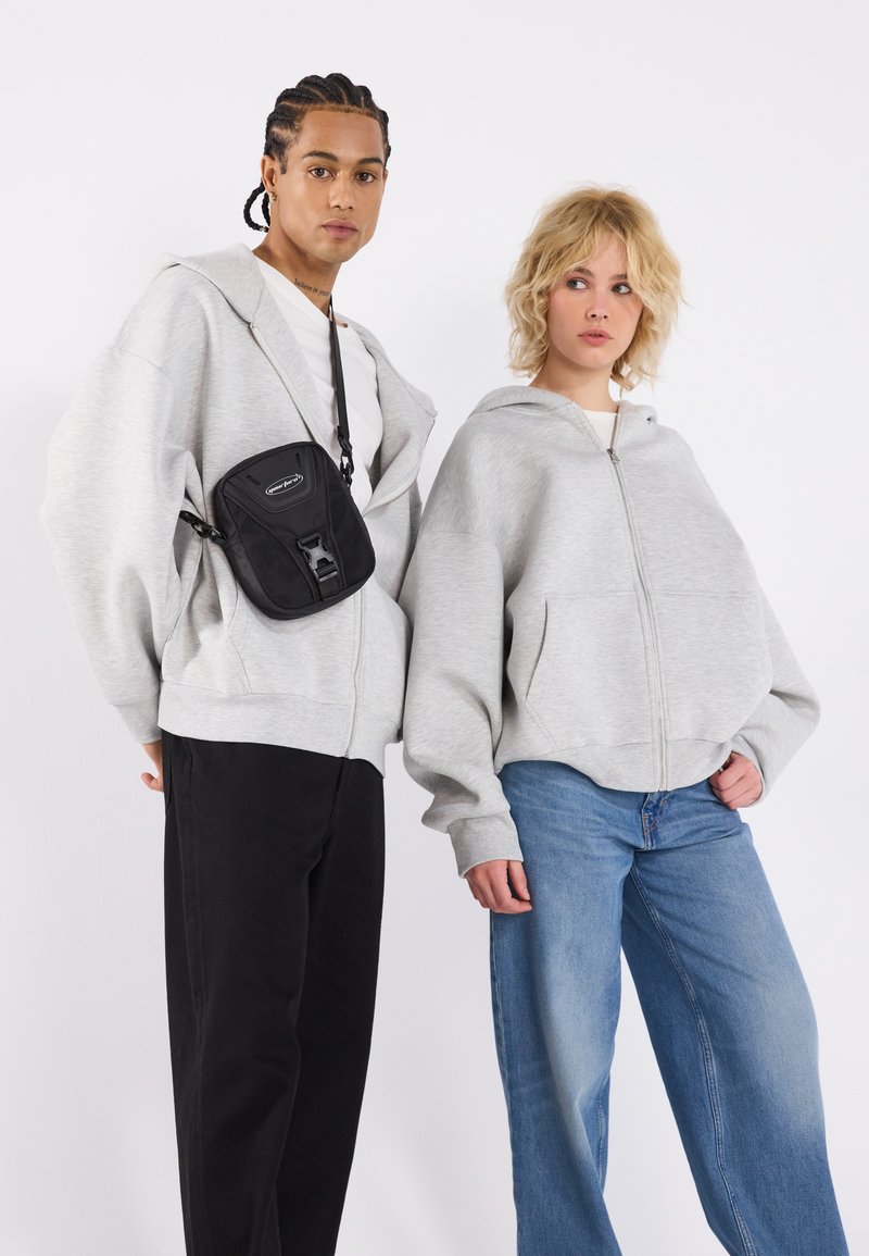 Sudaderas gris con cremallera y grandes bolsillos, usadas sobre una camiseta blanca, combinadas con jeans negros y azules. Incluye un pequeño bolso bandolera negro.
