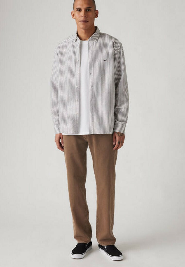 Man wearing a white t-shirt, gray striped button-up shirt, brown pants, and black slip-on shoes standing against a plain background.