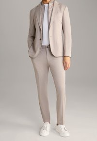 Ljust beige kostym med skräddarsydd kavaj och smala byxor, i kombination med en vit skjorta och vita sneakers. Slät tygtextur, minimal mönstring.