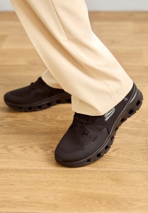 Person iført beige bukser og sorte Skechers-sneakers træder på en lys trægulv, der viser detaljer af skosålen.