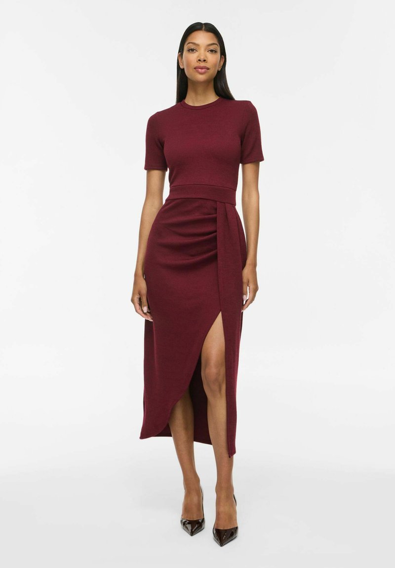 Femme debout en robe midi bordeaux ajustée à manches courtes avec fente devant et escarpins noirs, sur fond blanc uni.