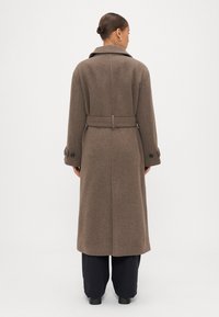 Brūns vilnas mētelis ar jostu viduklī, garām piedurknēm un pogātām aprocēm. Taisnas grieztas, ar aizmugures venti, paredzēts siltumam un aizsardzībai.