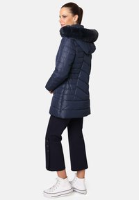Chaqueta de plumas azul marino con capucha y borde de piel sintética, patrón acolchado, cierre de cremallera, combinada con pantalones recortados azul marino y zapatillas deportivas blancas de caña alta.