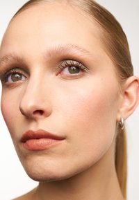 Portrait en gros plan avec une peau lisse, des lèvres brun clair, un maquillage des yeux subtil et des yeux noisette. Une simple boucle d'oreille en argent met en valeur le look.