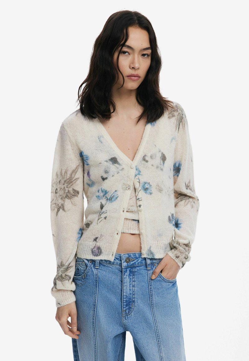 Cardigan en tricot crème avec un col en V, des manches longues et des motifs floraux en bleu et gris. Associé à un jean taille haute bleu clair.