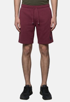 Uomo che indossa pantaloncini bordeaux con coulisse e scarpe da ginnastica nere, in piedi con le braccia rilassate lungo i fianchi su uno sfondo uniforme.