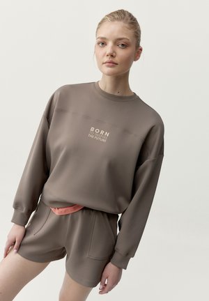 Mladá žena v hnedej súprave, mikina a šortky, s nápisom „BORN TO INSPIRE THE FUTURE“ na hrudi, stojaca pred jednoduchým pozadím.