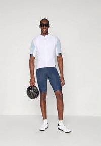 Homme en maillot de cyclisme blanc et bleu clair, shorts bleu marine, chaussures de cyclisme blanches, lunettes de soleil, tenant un casque noir, se tenant devant un fond uni.