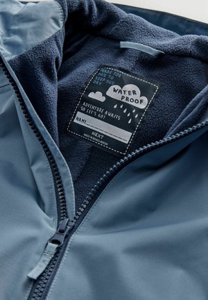 Vesimproof takki, jossa on sininen ulkopinta ja pehmeä tummansinisestä fleece-materiaalista vuori. Varustettu edessä vetoketjulla ja kankaiden hoitoetiketillä, jossa on leikkisiä kuvastoja.