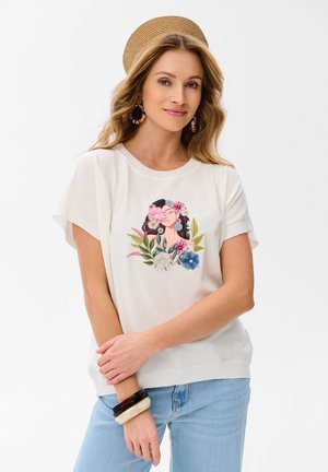 Kobieta w beżowym kapeluszu i białym topie z krótkim rękawem z kwiatowym haftem portretowym, jasnoniebieskich dżinsach, kolczykach kołach i bransoletkach, lekko się uśmiechająca.