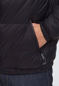 Persona con la mano destra nella tasca con zip di un piumino nero, che indossa jeans blu scuro, mostrata dalla vita a metà busto.