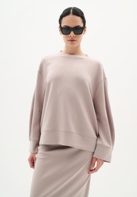 Överdimensionerad beige sweatshirt med rundad halsringning och sänkta axlar. Slät textur, ihop med en matchande beige kjol. Svarta rektangulära solglasögon.