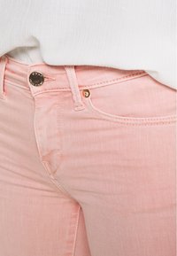 Vaaleanpunaiset farkut, joissa on hienovarainen tekstuuri, kullanväriset yksityiskohdat napissa ja taskuissa sekä ommellut yksityiskohdat saumoissa.