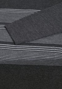 Pull en tricot gris foncé avec des rayures blanches et grises horizontales, montrant un gros plan de la manche et de la zone de la poitrine.