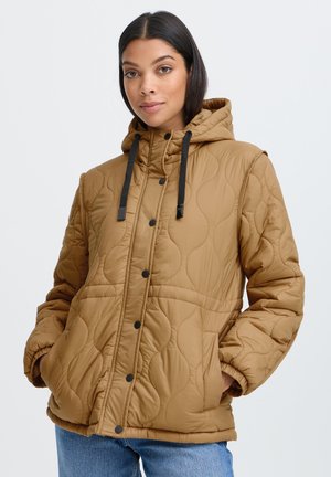 Femme aux cheveux foncés portant une veste matelassée beige à capuche avec des boutons noirs et les mains dans les poches, debout devant un fond clair uni.