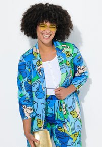 Ensemble de blazer et pantalon multicolore présentant un art linéaire abstrait en bleu, vert et beige. Accompagné d'une pochette dorée et de lunettes de soleil jaunes.