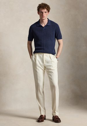 Άνδρας στέκεται με τα χέρια στις τσέπες, φοράει μπλε σκούρο κοντομάνικο πόλο, εκρού πιετέ παντελόνι, καφέ μοκασίνια με φούντα και ρολόι χειρός σε ουδέτερο φόντο.