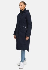 Marineblauer langer Mantel mit Kapuze, versehen mit einem Reißverschluss vorne, seitlichen Taschen und Knopfdetails; getragen über hellen Jeans und weißen Sneakers.