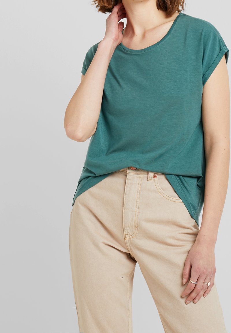 Femme portant un haut à manches courtes bleu sarcelle rentré dans un pantalon beige taille haute, avec une main touchant le cou et des bagues aux doigts.