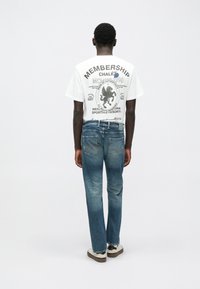 Άτομο που στέκεται με την πλάτη, φο ράει λευκό γραφικό T-shirt, μπλε τζιν με πίσω τσέπες και λευκά αθλητικά παπούτσια με μαύρες λεπτομέρειες.