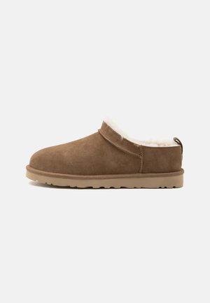 Zapatilla marrón de ante sin cierre con suela beige y forro de forro polar blanco, diseñada para calor y comodidad en interiores.