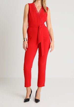 Femme portant une combinaison portefeuille rouge sans manches avec un bord en dentelle, une taille nouée, un pantalon longueur cheville, des talons noirs et un bracelet manchette doré.