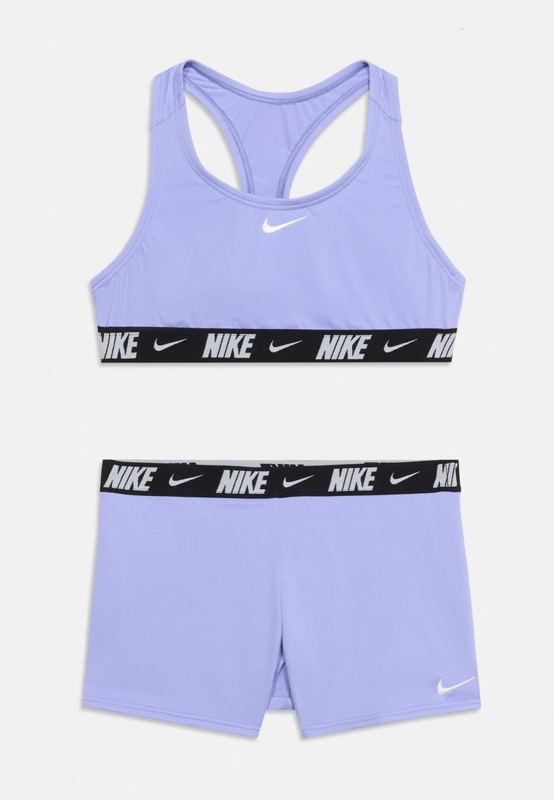 Ensemble brassière de sport et short Nike lilas avec bandes élastiques noires comportant des inscriptions blanches "Nike" et des logos swoosh sur un fond blanc.