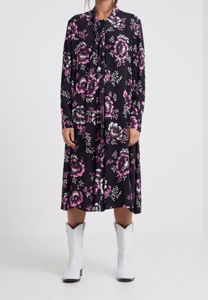 Das Blumenkleid aus dunklem Stoff zeigt rosa und weiße Blumen, lange Ärmel und eine Schleife am Kragen. Kombiniert mit weißen Cowboy-Stiefeln.
